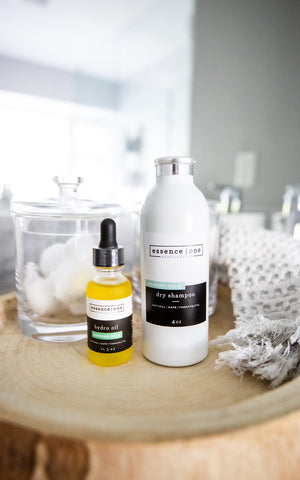 White bottle of Dry Shampoo | Essence One placed next to a yellow dropper bottle of hydro oil on a wooden surface with cotton balls and a textured white cloth in the background.