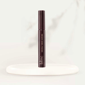 Sleek dark brown tube of Original Tubing Mascara | Blinc standing upright on a smooth white oval platform against a soft neutral background.