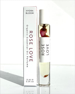 Glass roll-on bottle with visible rose petals containing Rose Love Organic Perfume Roll-on | Hydra Bloom, displayed next to its white rectangular packaging box.