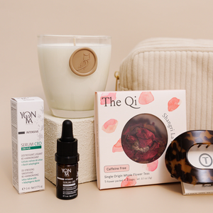A calming arrangement featuring Luxe Bath Ritual Kit | Live Love Spa with a CBD serum dropper bottle, a floral whole flower tea box, a lit candle in a glass jar, and a tortoiseshell wellness device on a neutral background.