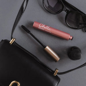Good Drama Mascara | Chella with black brush applicator displayed next to a black handbag and dark sunglasses on a gray surface.