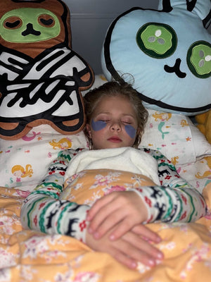 Child resting peacefully with eye patches on, lying in bed surrounded by two large Spa Bunny Plushie | Lucky Owl cushions featuring playful facial designs.