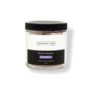 Clear glass jar with black lid containing lavender scented Shower Steamer | Essence One, labeled as natural, safe, and therapeutic, for body care and wellness.
