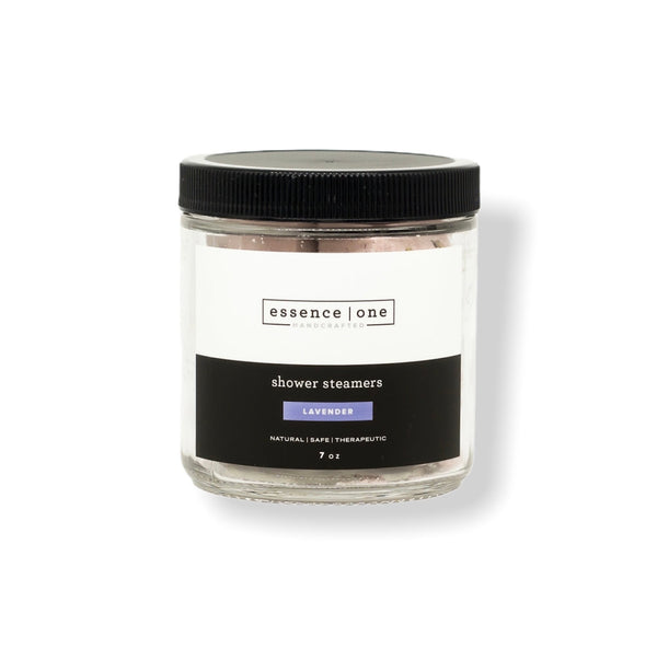 Clear glass jar with black lid containing lavender scented Shower Steamer | Essence One, labeled as natural, safe, and therapeutic, for body care and wellness.