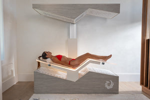 A woman in a red bikini lying inside a modern wellness device called Ammortal Chamber | Ammortal, designed for relaxation and recovery in a minimalistic room.