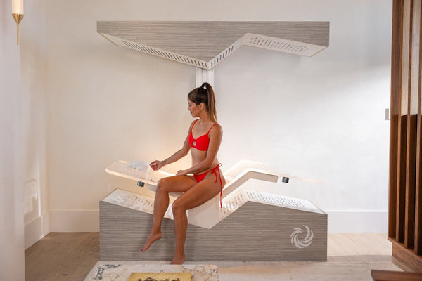 A woman in a red swimsuit sits inside the Ammortal Chamber | Ammortal wellness device, adjusting controls while relaxing in a modern, minimalistic room.