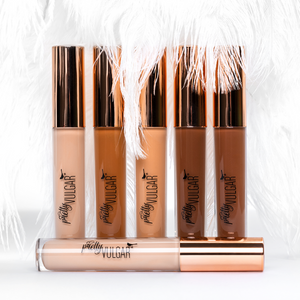 A range of concealer tubes in various skin tones with rose gold caps, featuring the product name Under Cover Concealer | Pretty Vulgar, displayed against a soft white feather background.