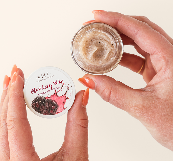 Hands holding an open jar of Blackberry Wine Lip Polish | Farmhouse Fresh showing the textured sugar lip polish with blackberry and wine graphics on the lid for skincare and lip care.