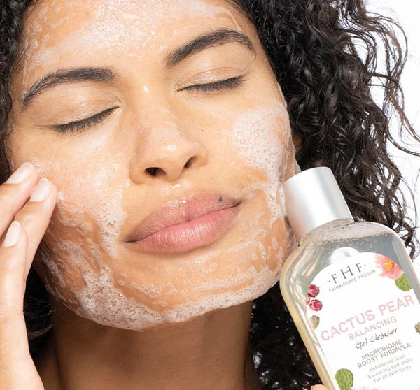 Woman gently cleansing her face with lather while holding a bottle of Cactus Pear Balancing Gel Cleanser | Farmhouse Fresh, showcasing a skincare routine.