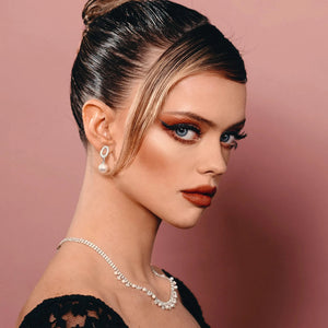 Close-up portrait of a woman with elegant makeup featuring dramatic lashes enhanced by Good Drama Mascara | Chella, wearing pearl earrings and a jeweled necklace against a soft pink background.