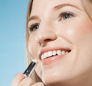 A close-up of a woman smiling while applying Coconut Beach® Hydrating Lip Balm | Farmhouse Fresh to her lips, showcasing skincare and lip hydration.