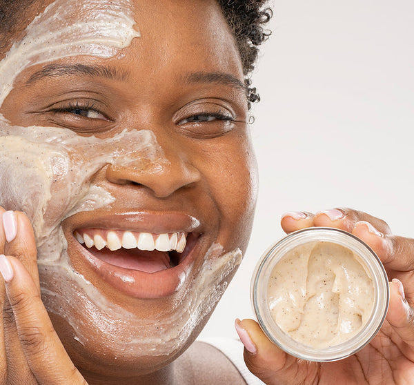 Smiling person with a creamy exfoliating scrub applied on their face holding an open jar of Finely Awake Plantfoliant Silkening Face Cleansing Polish | Farmhouse Fresh, highlighting skincare and self-care.
