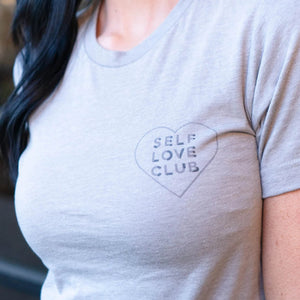 Close-up of a person wearing a light gray Self Love Club Unisex Crew Neck T-Shirt | Lucky Owl featuring a small heart graphic with the words "SELF LOVE CLUB" on the chest.