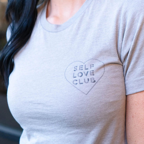 Close-up of a person wearing a light gray Self Love Club Unisex Crew Neck T-Shirt | Lucky Owl featuring a small heart graphic with the words "SELF LOVE CLUB" on the chest.
