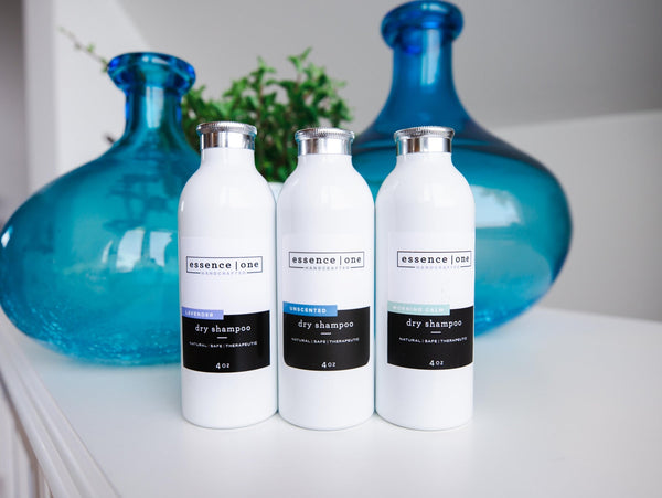 Three white bottles of Dry Shampoo | Essence One with silver caps displayed on a white surface, set against a background featuring two large blue glass vases and greenery.