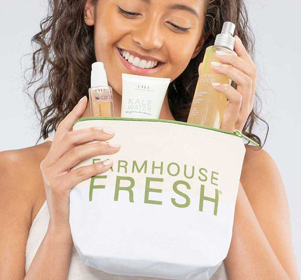 A smiling woman holding the FHF Canvas Cosmetic Bag | Farmhouse Fresh filled with skincare products including a spray bottle, serum, and hydrating cleanser.
