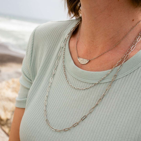 Close-up of a person wearing layered silver necklaces featuring the Frolic Paperclip Necklace /silver tone | Purpose Jewelry over a light green ribbed top, with a blurred beach background.