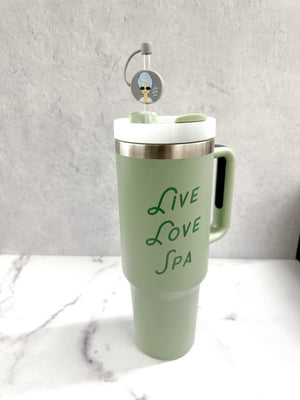 A pale green travel tumbler with a handle, featuring the words "Live Love Spa" in elegant green script and a straw topped with the Spa Girl Straw Topper | Live Love Spa accessory.