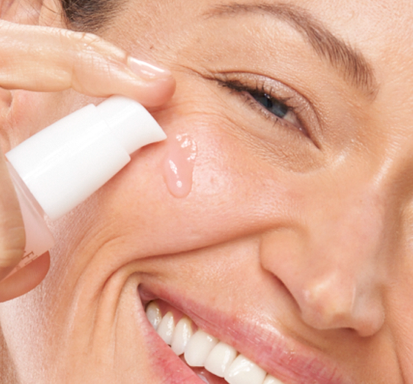 Close-up of a smiling woman applying a light pink serum to her cheek using a white pump bottle, showcasing skincare with Lustre Rose™ Serum-in-Oil | Farmhouse Fresh.