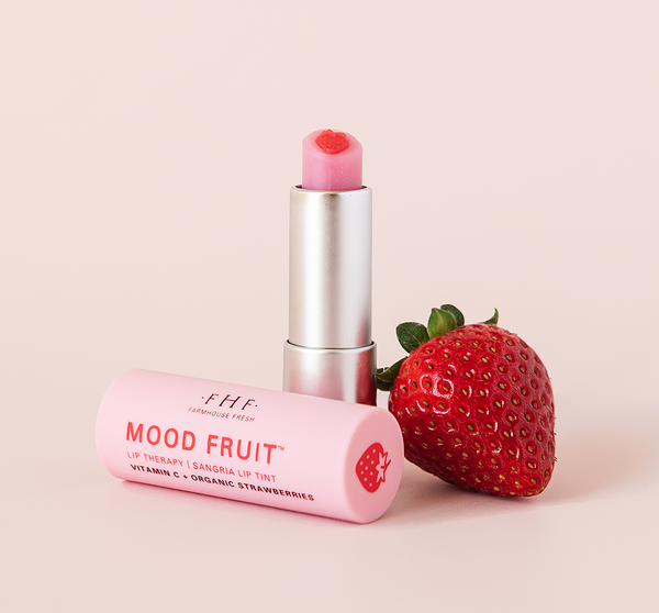 Strawberry Mood Fruit™ Lip Therapy | Farmhouse Fresh in a pink tube with a heart-shaped balm top, displayed next to a fresh strawberry, highlighting skincare and lip care.
