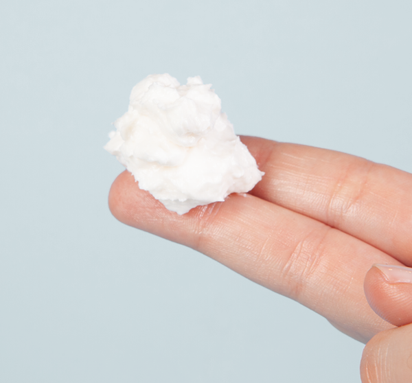 Close-up of a generous dollop of Moon Dip Back To Youth Ageless Body Mousse | Farmhouse Fresh held on two fingertips against a light blue background, showcasing its creamy skincare texture.
