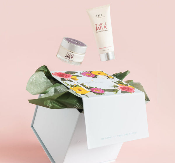 Open white gift box with floral design and green tissue paper, featuring skincare products from Silky, Milky & Most Loved Naked Nourishment Set | Farmhouse Fresh floating above on a pink background