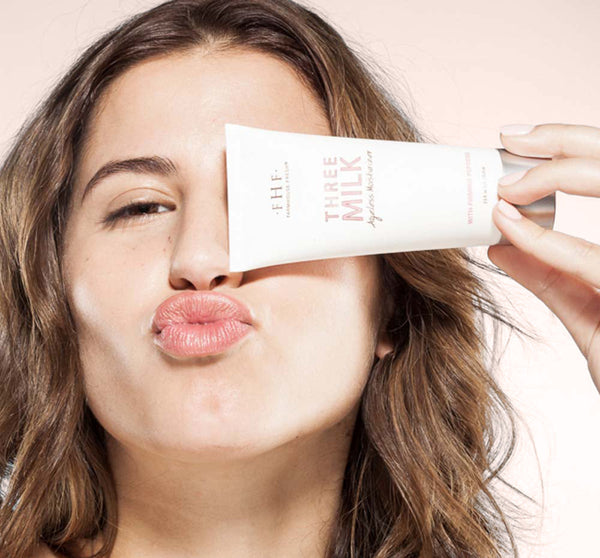 Young woman with clear skin playfully holding a skincare tube close to her face, promoting the Silky, Milky & Most Loved Naked Nourishment Set | Farmhouse Fresh body care collection.