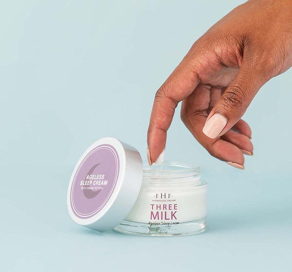 Hand dipping finger into an open jar of Three Milk Ageless Sleep Cream on a blue background, representing skincare from the Silky, Milky & Most Loved Naked Nourishment Set | Farmhouse Fresh.
