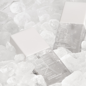 Two clear square bottles with white caps labeled Face Tan Water - Dark | NUDA surrounded by ice cubes, highlighting skincare and refreshing body care product.