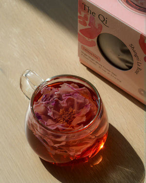 Clear glass cup filled with rose-infused herbal tea next to The Qi Shangri La Rose box, showcasing a calming and luxurious skincare moment from Luxe Bath Ritual Kit | Live Love Spa.