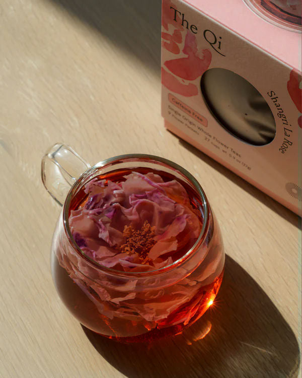 Clear glass cup filled with rose-infused herbal tea next to The Qi Shangri La Rose box, showcasing a calming and luxurious skincare moment from Luxe Bath Ritual Kit | Live Love Spa.