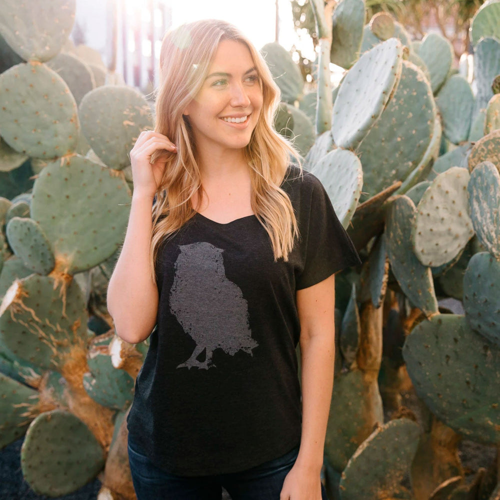 Woman wearing a black Distressed Owl Women's T-Shirt | Lucky Owl with a faded owl graphic, standing outdoors surrounded by green cactus plants, smiling and looking to the side