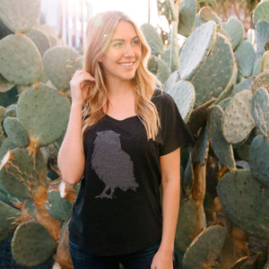 Woman wearing a black Distressed Owl Women's T-Shirt | Lucky Owl with a faded owl graphic, standing outdoors surrounded by green cactus plants, smiling and looking to the side