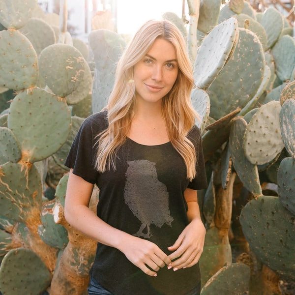 Woman standing in front of cactus plants wearing a black Distressed Owl Women's T-Shirt | Lucky Owl with a subtle owl graphic on the front, smiling with hands lightly clasped.