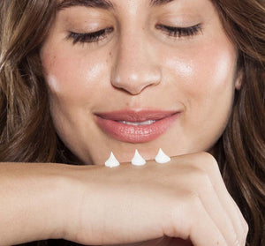 A woman smiling gently with eyes closed, applying three small dollops of Pink Moon Shea Butter Hand Cream | FarmHouse Fresh on the back of her hand, showcasing smooth glowing skin.