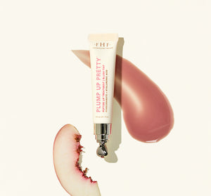 Plump Up Pretty Peptide Lip Treatment | Farmhouse Fresh tube with a metal applicator, placed next to a smear of rosy lip treatment and a bitten peach slice on a light background.