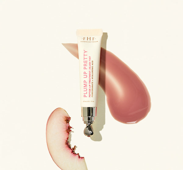 Plump Up Pretty Peptide Lip Treatment | Farmhouse Fresh tube with a metal applicator, placed next to a smear of rosy lip treatment and a bitten peach slice on a light background.