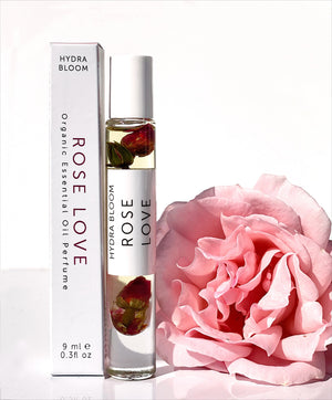 Rose Love Organic Perfume Roll-on | Hydra Bloom next to a soft pink rose, featuring a clear glass bottle with rose petals inside and a white box packaging, showcasing a natural skincare product.