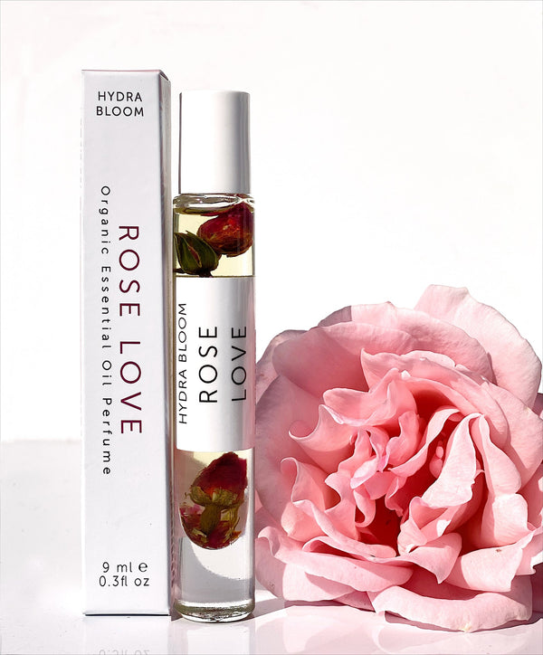 Rose Love Organic Perfume Roll-on | Hydra Bloom next to a soft pink rose, featuring a clear glass bottle with rose petals inside and a white box packaging, showcasing a natural skincare product.