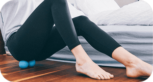Person using RAD Roller Stiff | RAD, a blue recovery tool placed under their lower back while sitting on a wooden floor, demonstrating muscle release and body care.