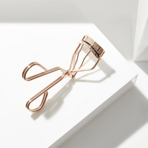 Procurl Lash Curler | Tweezerman in a metallic rose gold finish placed on a white geometric surface with soft natural lighting highlighting the sleek design.