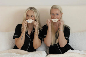 Two women sitting in bed wearing black pajamas, holding white strips of Mouth Tape | Seald™ Wellness over their mouths, promoting nighttime wellness and restful sleep.