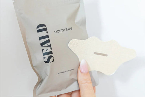 Hand holding a single piece of Mouth Tape | Seald™ Wellness in front of a beige resealable package with the brand name Seald visible on it.