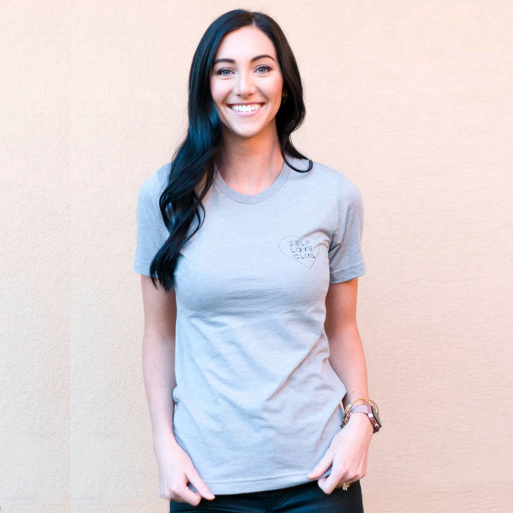 A smiling woman with long dark hair wearing a light gray Self Love Club Unisex Crew Neck T-Shirt | Lucky Owl, standing against a plain light peach background.