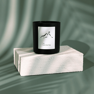 Black candle with minimalistic white label depicting a yoga pose, placed on a textured white block against a soft green background, featuring No. 1 Sunrise Island Yoga (Bali) Candle | Lucky Owl.