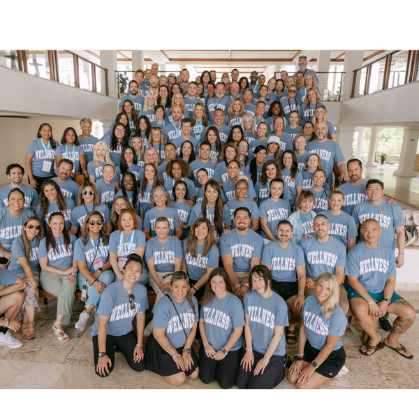 Large diverse group of people wearing matching blue Wellness Varsity T-Shirt | Lucky Owl, gathered indoors smiling and posing together for a group photo.