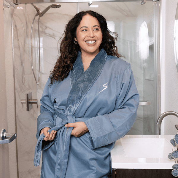 Woman smiling wearing a Microfiber Shimmer Lined Robe - Teal | Mansfield standing in a modern bathroom near a sink and shower.