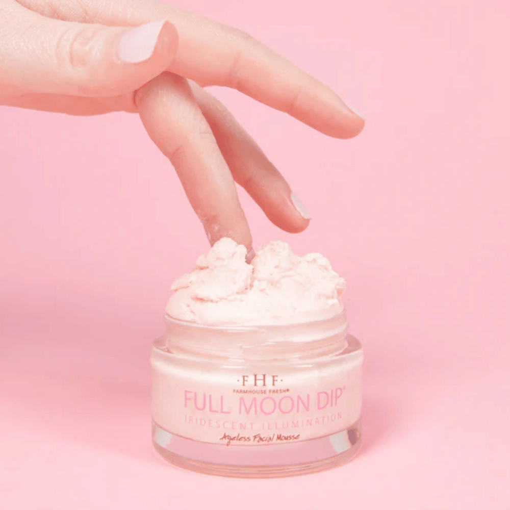 A hand dipping a finger into a jar of Full Moon Dip® Iridescent Illumination Ageless Facial Mousse with Peptides + Retinol | Farmhouse Fresh against a soft pink background.
