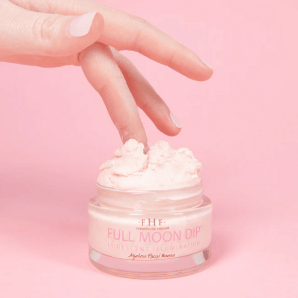 A hand dipping a finger into a jar of Full Moon Dip® Iridescent Illumination Ageless Facial Mousse with Peptides + Retinol | Farmhouse Fresh against a soft pink background.