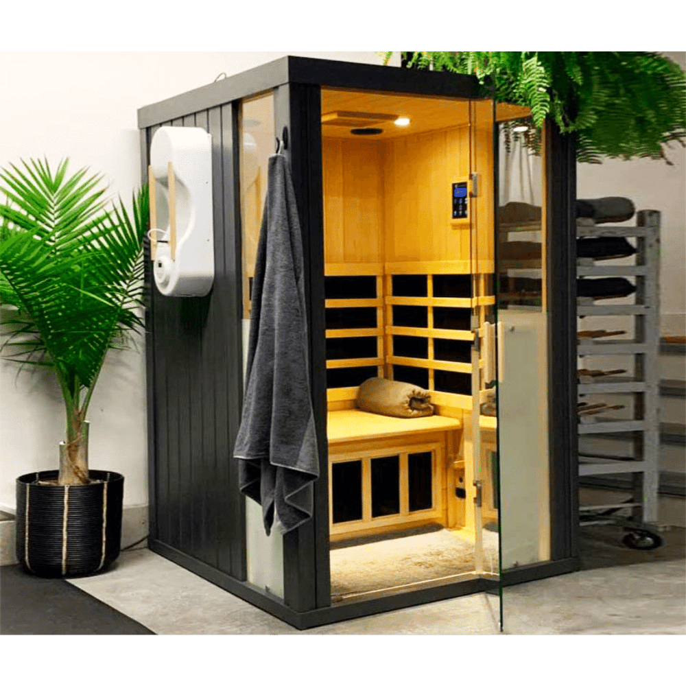 Modern black and wood HaloSauna™ | Halotherapy Solutions unit with glass door, interior seating, digital control panel, and gray towel hanging outside, placed next to a green plant for wellness and skincare.